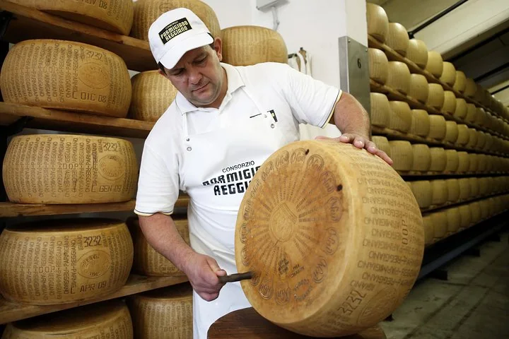 A cheesemaker in a Parmigiano Reggiano apron tapping a whole wheel with a small hammer in an aging room