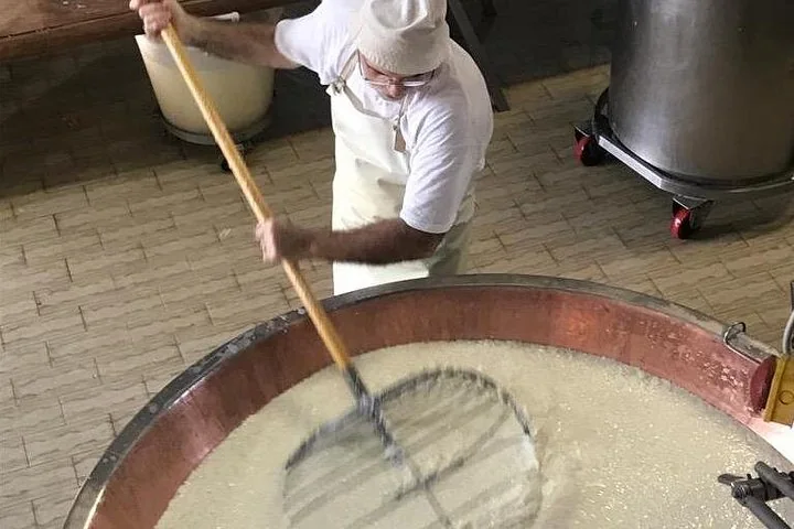 A cheesemaker stirring milk in a copper vat with a large wire whisk on a long wooden pole