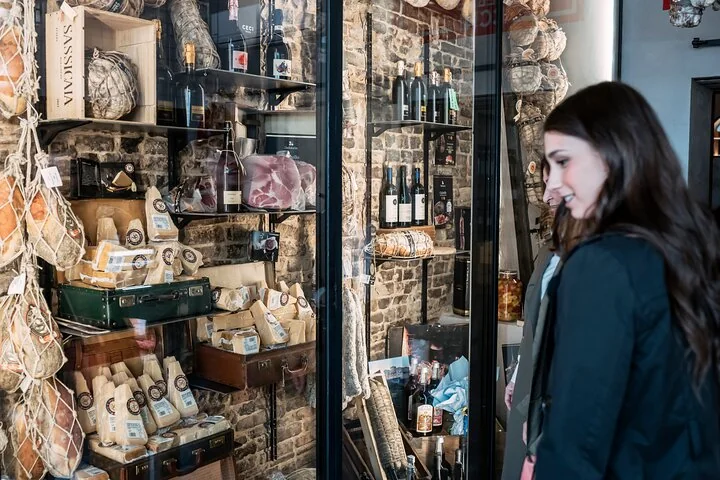 A woman looking into a deli packed with wedges of Parmigiano, hams, salami and bottles of wine
