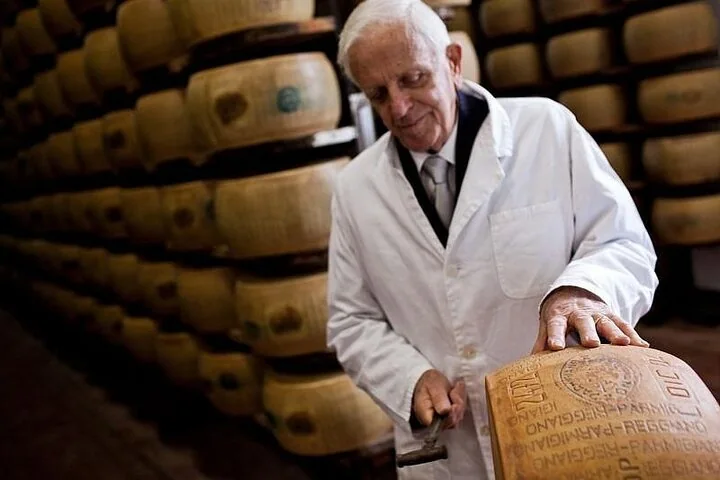 An elderly expert in a white coat tapping a Parmigiano wheel with a small hammer in a dim aging room