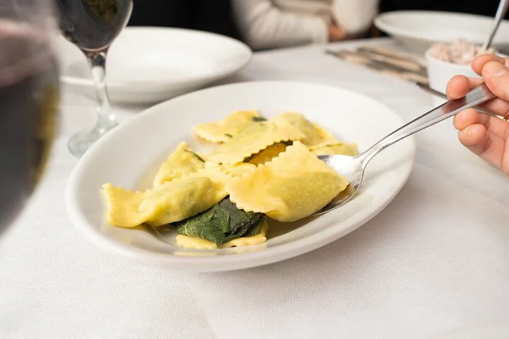 Square stuffed pasta with a green leaf on a white plate, with a fork and a glass of red wine on a white tablecloth