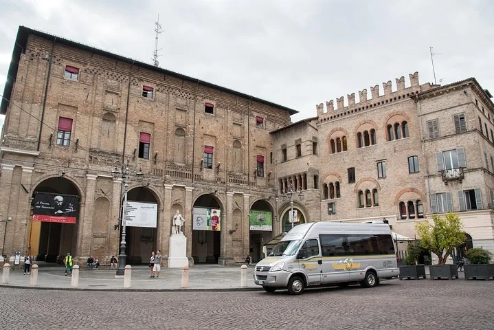 A white tour minibus parked on a cobbled square in central Parma in front of arcaded brick palazzi