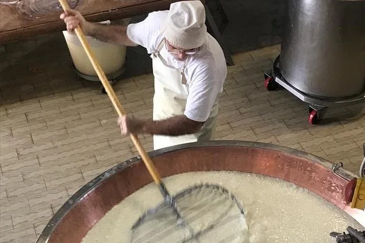 A cheesemaker stirring milk in a copper vat with a large wire whisk on a long wooden pole